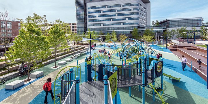 A distant view of an inclusive playground in bright blues and greens with a city surrounding it. 