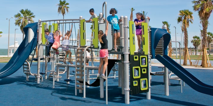 Children playing on a gray, navy and lime green play structure with beach and ocean in the back ground. 