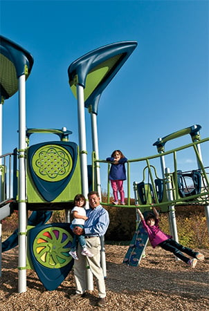 Steve King Landscape Structure founder holding a toddler in front of a PlayBooster playground.