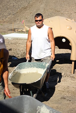 Volunteer pushing wheelbarrow with concrete at a playground building site.