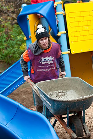 KaBOOM! volunteer pushing wheelbarrow with concrete at a playground building site.
