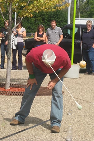 Landscape Structures employee playing a game where he has to knock a water bottle over with a tennis ball dangling from tights attached to his head.