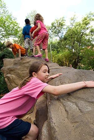 A young girl climbs up onto a Pinnacle rock climber where a group of children stand at the peak of the climber.