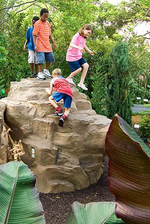 A group of children climbing on a Pinnacle rock climber surrounded by a garden.