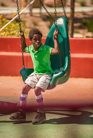 A boy sits on the edge of an accessible green swing 