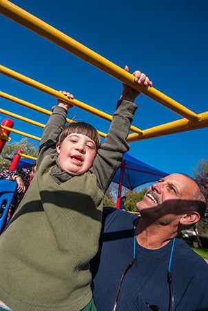 A boy smiles and a man helps him as he navigates an overhead monkeybar structure. 
