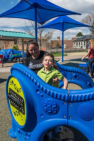 A boy stands inside a blue playground structure while a woman helps him from outside. 