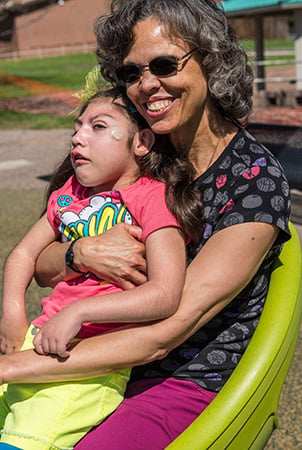A smiling woman sitting on an adaptive spinning seat holds a young girl. 