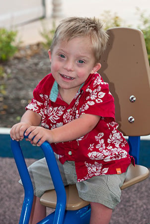 A little boy in a red Hawaiian shirt sits in a seat that's part of a playground component. 
