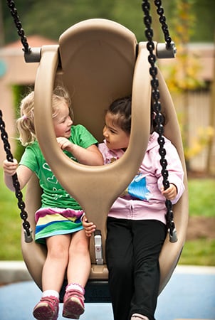 Two girls smile at each other as they swing in an adaptive swing. 