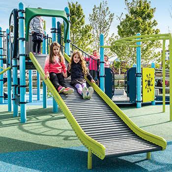 Two girls slide down a Rollerslide, which is an inclusive commercial playground slide
