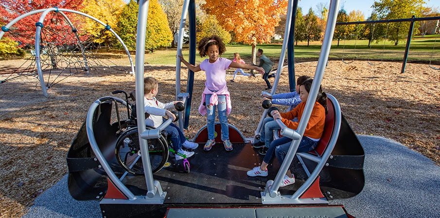 Four children playing on a We-Go-Swing, one of whom is in a wheelchair. Fall foliage in background.