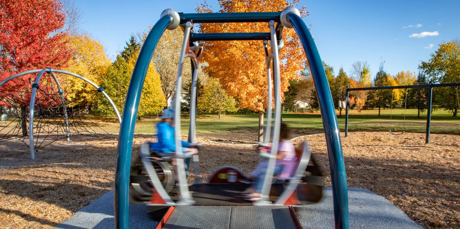 Two children swinging on the We-Go-Swing handicap swing for playgrounds, and one is in a wheelchair. The image is blurred to show the speed of the children swinging.