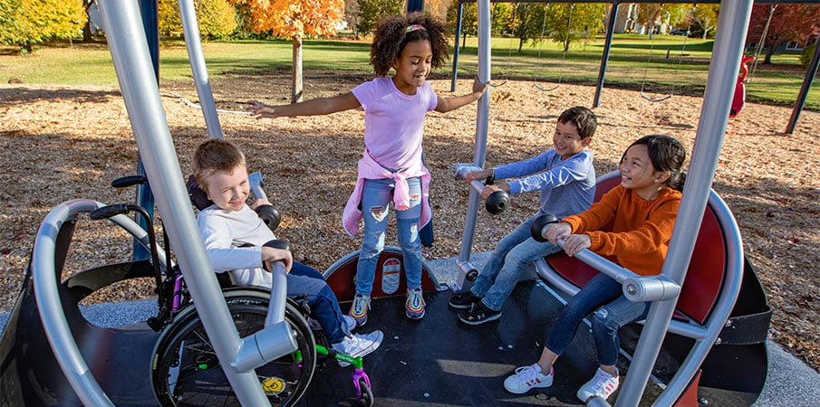 Four children playing on the We-Go-Swing wheelchair-accessible swing for playgrounds. One girl is standing, while two are sitting on a seat and another child is in a wheelchair.