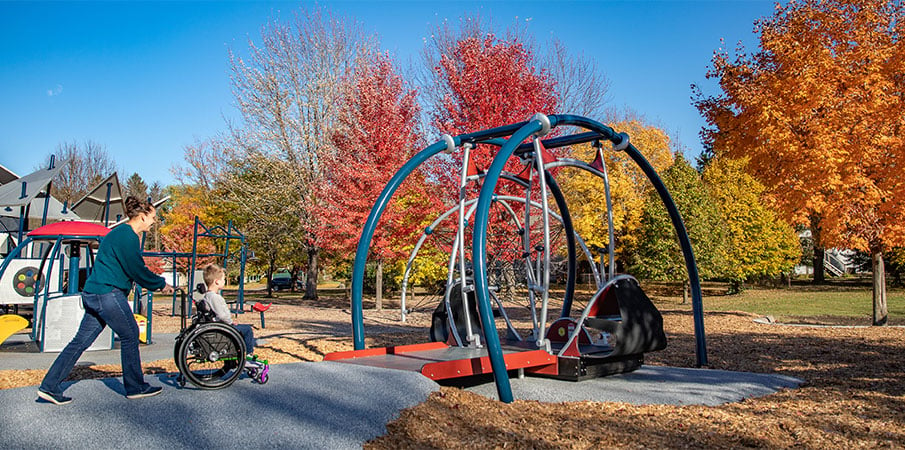 An adult pushes a child in a wheelchair up a ramp toward the We-Go-Swing, playground swing for wheelchair users