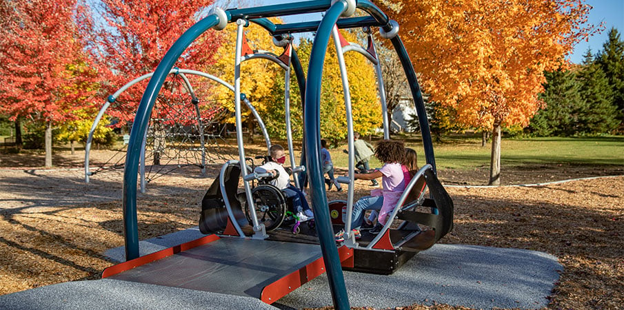 Three children playing in the first inclusive and wheelchair-accessible playground swing, the We-Go-Swing. One child is in a wheelchair.