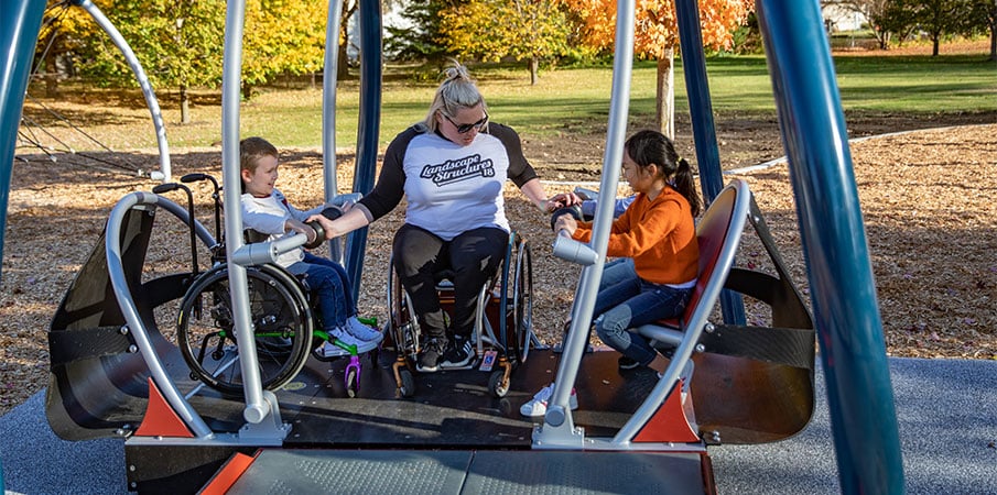 Inclusive play expert Jill Moore sits in the We-Go-Swing with two children, one of whom is in a wheelchair.