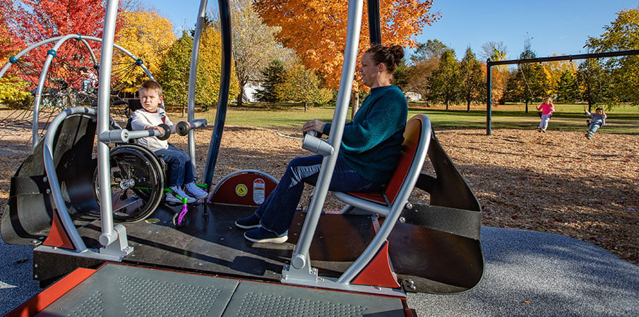 An adult and a child sit in the We-Go-Swing, a handicap-accessible swing for commercial playgrounds. The child is in a wheelchair.