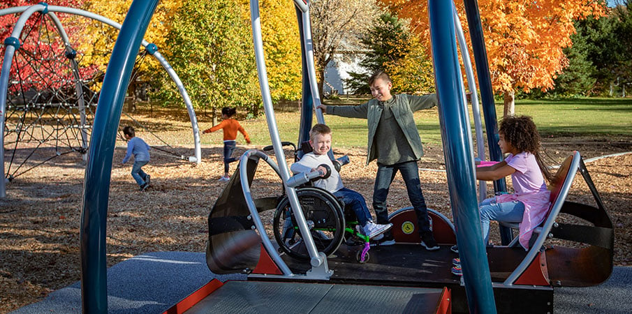 Four children play in the inclusive We-Go-Swing, which is a wheelchair-accessible playground swing set. One boy stands in the middle, two girls sit on one side, and a child in a wheelchair sits on the other side.