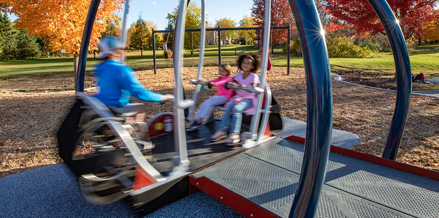 Two children and an adult in a playground wheelchair swing in the handicap-accessible We-Go-Swing. The image is blurred to show the speed of swinging.