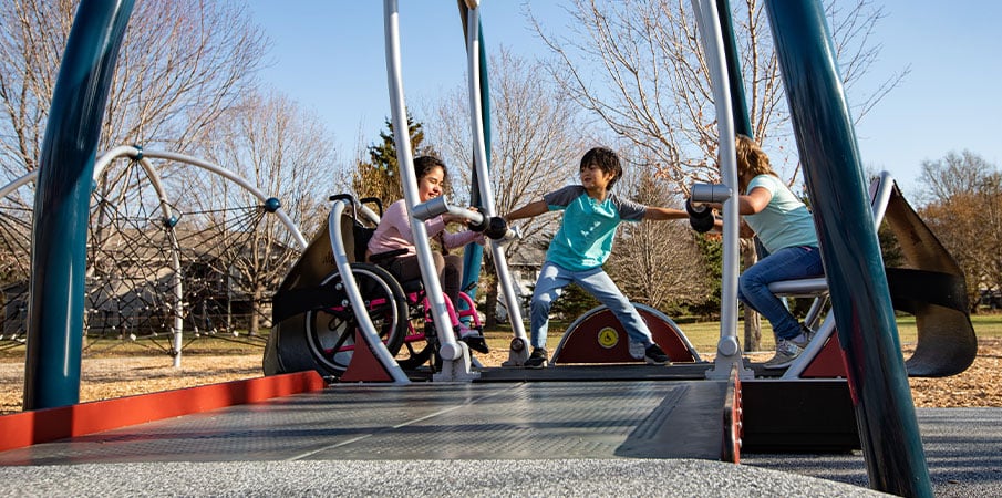 Three children playing in We-Go-Swing with one girl in wheelchair. Boy has his arms stretched from one side of swing to the other.