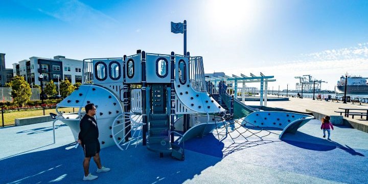 Playground with shiny silver climber that looks like a ship and blue surfacing with apartment buildings and a waterfront in the background. 