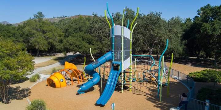 Overhead image of a playground with a tall blue and gray climber with reed-like posts and an orange goldfish climber with chipped wood surfacing surrounded by trees. 