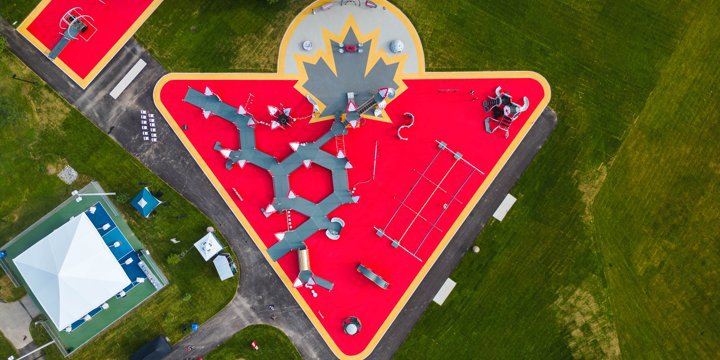 Overhead view of playground with red surfacing shaped like an upside-down triangle and a gray maple leaf motif at the top. Playground shows lots of gray ramping throughout. 