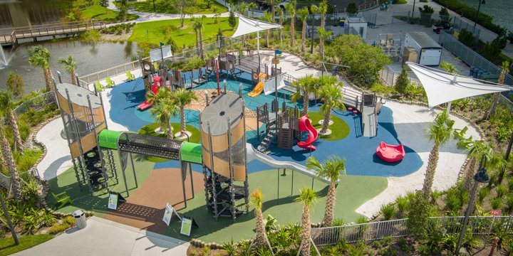 A large playground surrounded by palm trees with two tall climbing towers in the front connected by a crawl tunnel and multiple red slides, a red spinner and several climbing structures behind. 