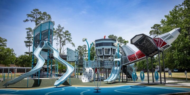 Space themed playground with a red and silver spaceship climber to the right, a space station-themed climber in the middle and a light blue and silver tower to the left with two slides. 