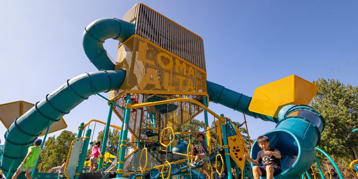Looking up at a towering play structure with "Loma Alta" machined into a perforated panel on the tower, with two turquoise blue twisty slides and an orange rope climber in the foreground. 
