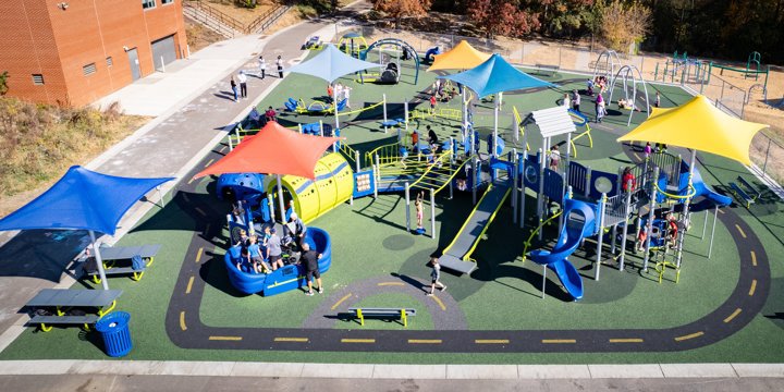 Expansive playground including custom surfacing with a "road" motif that runs around the play area. Multiple colorful shade sails cover complex, connected play structure that is dense with play features, including a sensory tunnel, roller slide and other inclusive elements. 