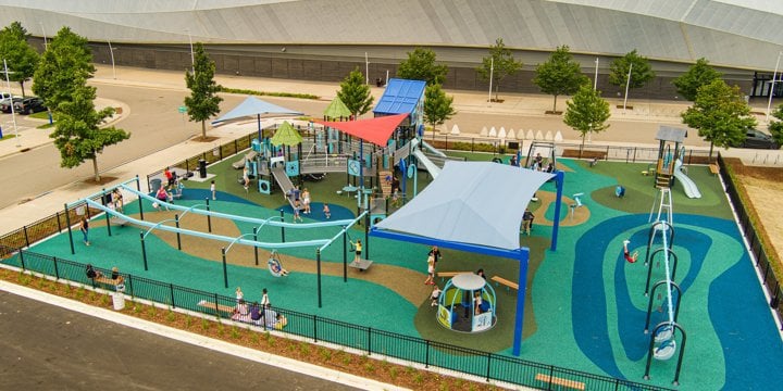Overhead view of playground with stadium in the background. A dual bay track ride in the front left and an accessible spinner in the foreground, mulitple bay swing set to the right and a multifaceted playstructure to the back left. 