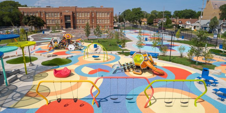 A very large playground with brighly colored surfacing and a three-bay geometric swing set in the foreground and two geometric play structures among other brightly colored play features. 