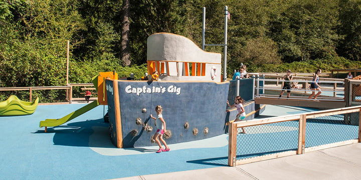 Girl runs in front of the prow of a blue and white custom concrete ship on the crescent creek playground. 