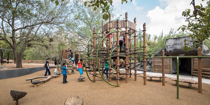 Brown and green, nature-themed playground structure with log benches that blends in with trees in the background and a light brown playground surface. 