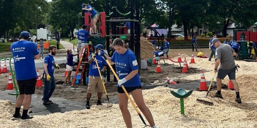 Adults in matching blue t-shirts rake a loose fill playground surface while others consult with each other. A playground is being constructed in the background.