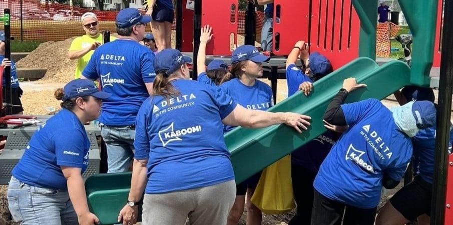 A group of women in matching blue t-shirts hold a playground slide and look toward where the slide connects to the deck, in the process of putting the pieces together.