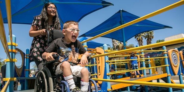 A boy in a wheelchair laughs as a girl pushes him across a playground ramp 