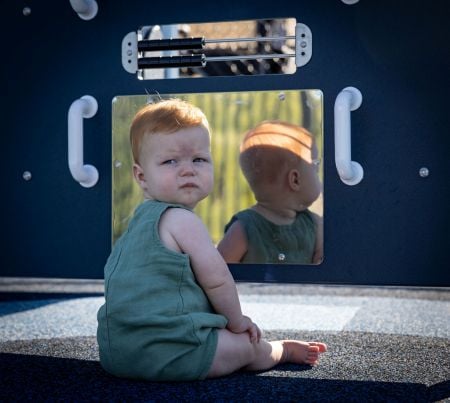 A toddler sits in front of a mirror panel on a playground and looks back at the camera