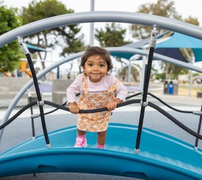 Little girl holds onto cable from playground bridge and smiles at camera