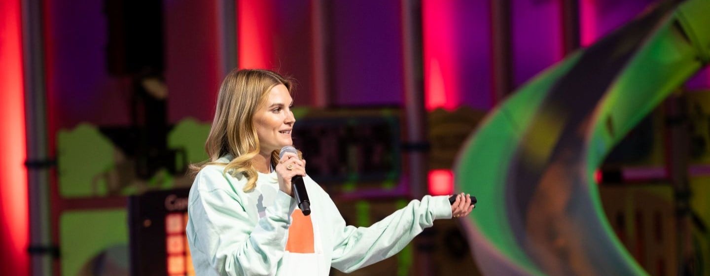Woman in blue sweatshirt is on a stage with playground equipment behind her speaks on a microphone.