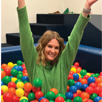 Woman sitting in a ball pit with hands raised, smiling