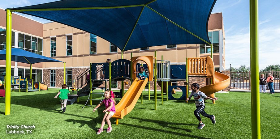 Playground at Trinity Church with children playing and large dark blue shade structure overhead.