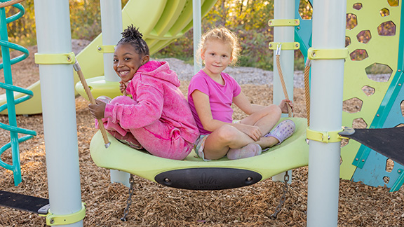 Two children sitting in a swing under a playground deck.
