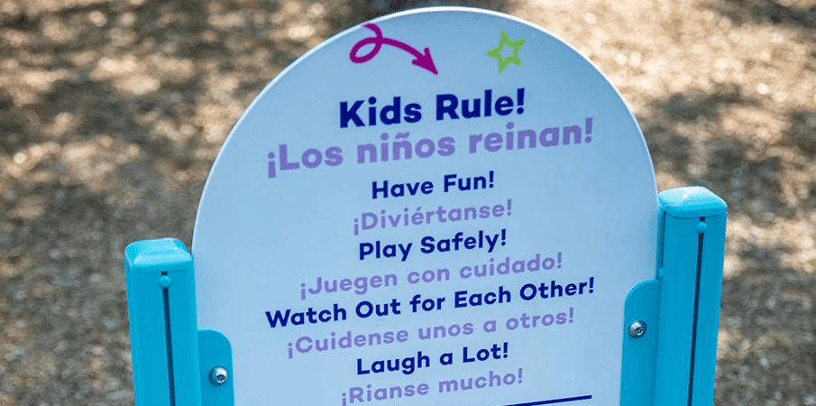 Closeup on a playground sign reading "Kids Rule" "los ninos reinan". It is a light blue sign that is rounded at the top with turquoise posts.