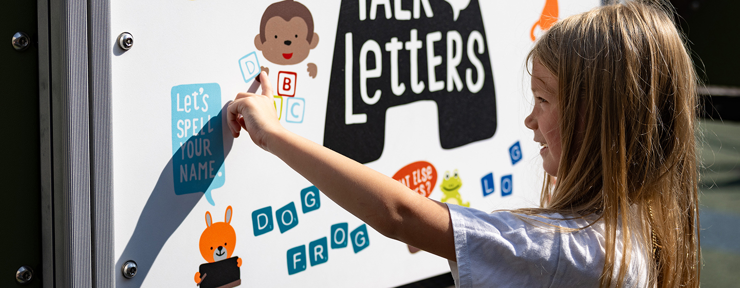 A girl points at a sign on a playground that has colorful graphics and words.