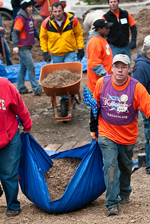 Two men pull a tarp filled with mulch. One of them wears a purple bib that reads "Kaboom!" 