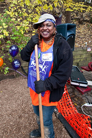 A woman smiles holding the handle of a rake. She is wearing a bib that reads "Kaboom!" 