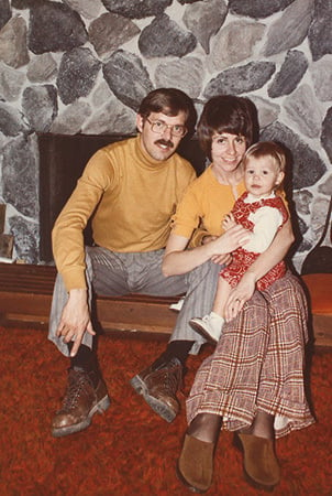 Barb and Steve King sitting on a fireplace hearth holding a child. 